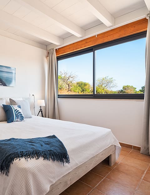 Dormitorio con paredes blancas, vigas de madera vistas, suelos de baldosas y cama frente a una ventana.
