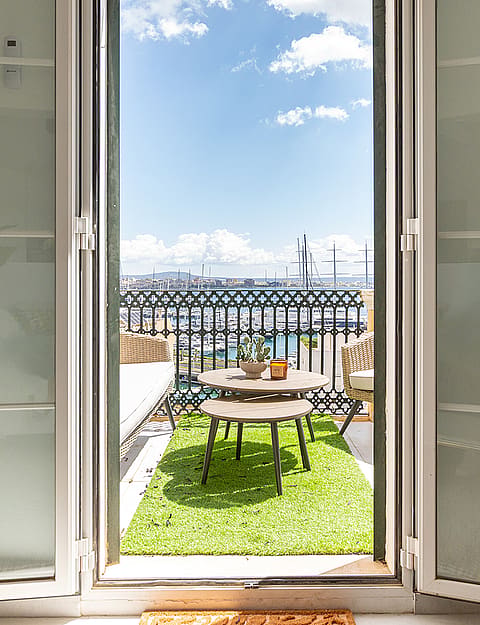 A view through white-framed glass doors onto a small balcony with a green carpet, a bistro table and chairs, and a harbor view.