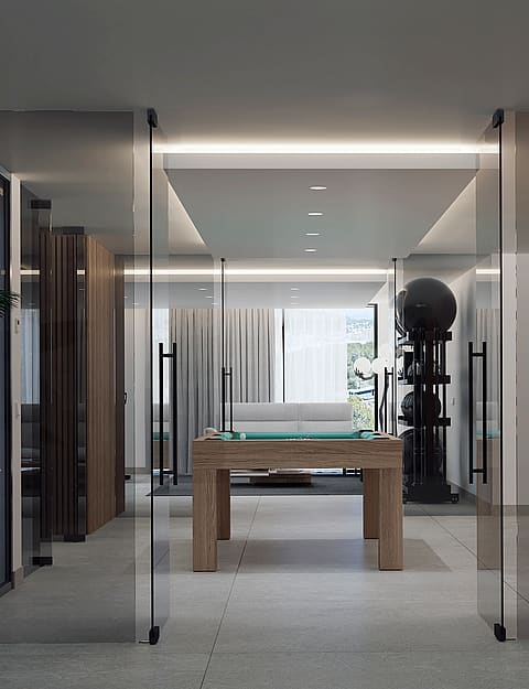 A modern games room featuring a wooden pool table and gym equipment, viewed through frameless glass pivot doors.