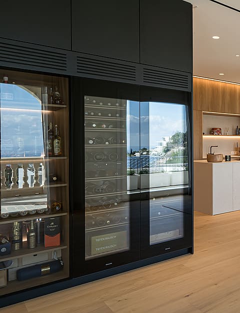 Open-plan dining area with a long wooden table, modern chandelier, and floor-to-ceiling glass wine fridge in a villa.