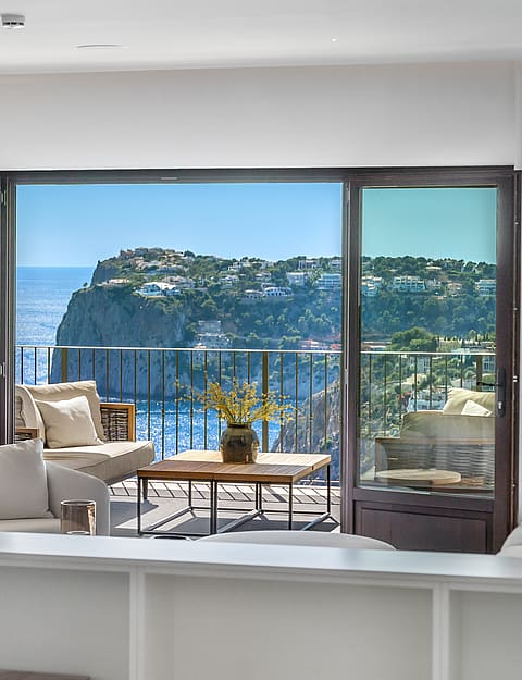 Interior view through dark-framed glass doors to a furnished terrace overlooking a coastal cliff and the blue sea.