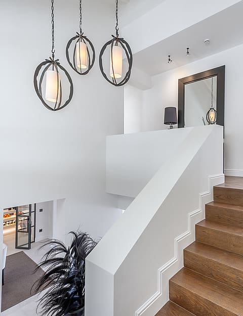 A white staircase with oak steps sits adjacent to a brushed steel elevator and three circular pendant lights.