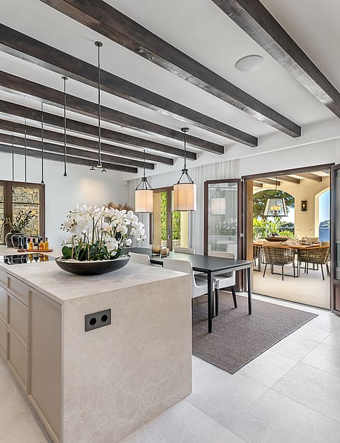 A wide stone kitchen island and dark ceiling beams in an open-plan room opening onto a sunlit outdoor dining terrace.