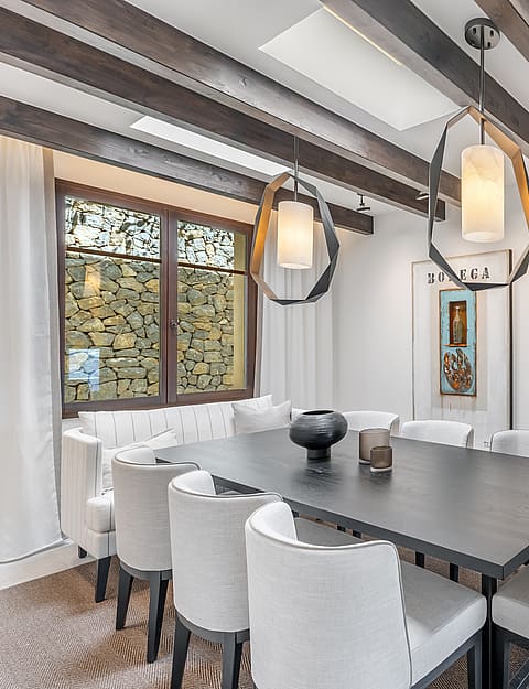 Modern dining room featuring a dark wood table, white upholstered chairs, exposed ceiling beams, and geometric pendants.