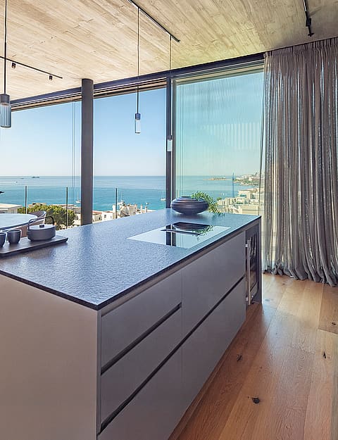 Modern kitchen island with dark stone countertop, light wood flooring, and floor-to-ceiling glass windows overlooking the sea.