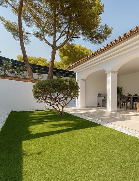 Covered outdoor terrace with white arched columns, dining table and wicker sofa opening onto lawn and mature pines