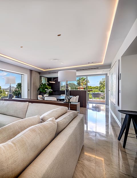 A contemporary open-plan living room featuring a large beige sectional sofa, polished stone floors, and floor-to-ceiling glass sliding doors opening to a lush garden view.