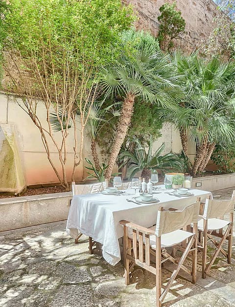 An outdoor dining table with white tablecloth and director chairs set on a gravel terrace shaded by mature trees.
