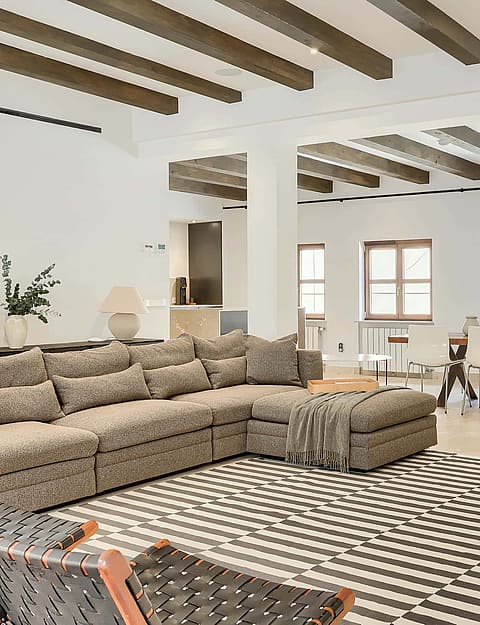 A spacious living room featuring a large patterned sofa, exposed dark wood ceiling beams, and a dining area in the background.