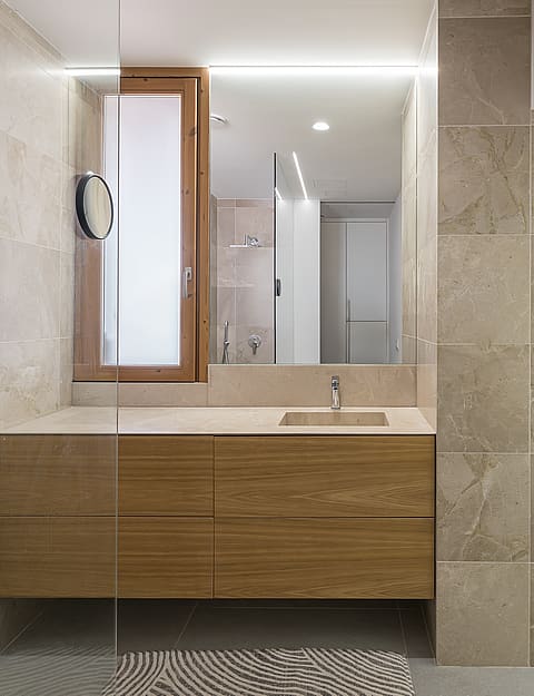 A modern bathroom featuring a wall-mounted wooden vanity, large rectangular mirror, and beige natural stone wall tiles.