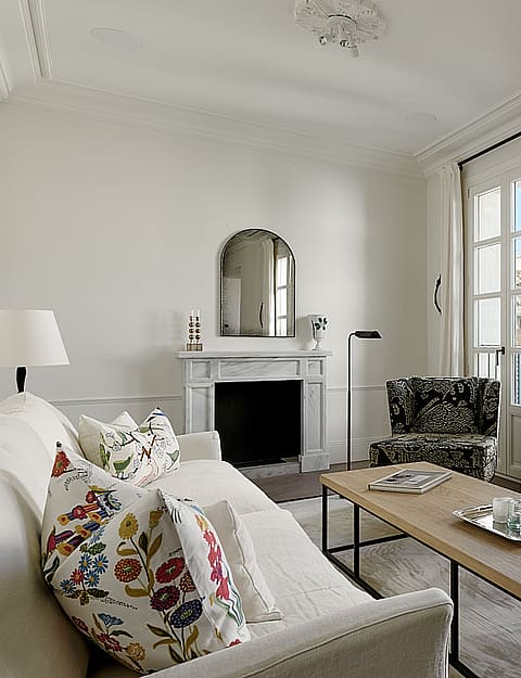 Bright living room with cream furnishings, wooden fireplace mantle, tall windows overlooking the city, and herringbone wooden floors