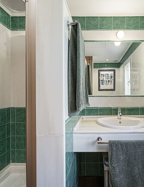 A bright bathroom with white and green square wall tiles, a glass-enclosed shower, a white vanity with a sink, and a rectangular mirror.