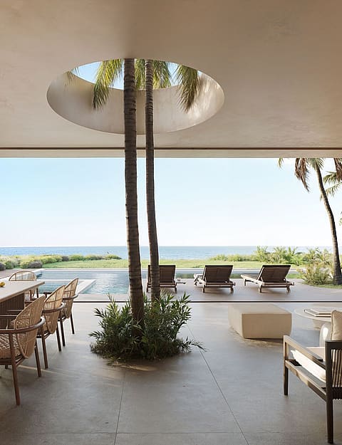 Outdoor dining and lounge area on a shaded terrace featuring a circular opening in the ceiling with a tree, overlooking a pool and the sea.