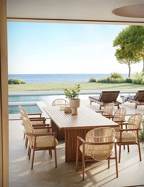 A slatted wood outdoor dining table with wicker chairs on a covered terrace, facing a swimming pool and the Mediterranean sea.