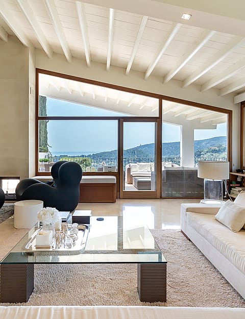 Spacious villa living room with white painted beams, fireplace, marble floors, designer seating and glazed wall with sea views
