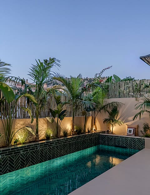 A rectangular outdoor swimming pool featuring dark green patterned tiles, integrated lighting, and tropical landscaping.