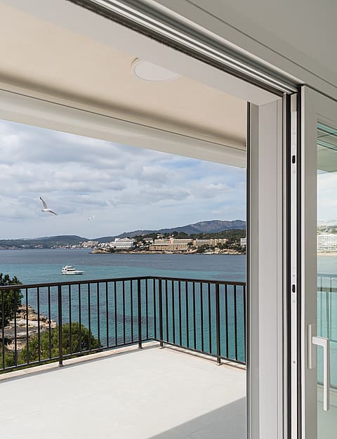 A white tiled terrace with dark metal railings overlooking a clear turquoise bay and distant mountains.