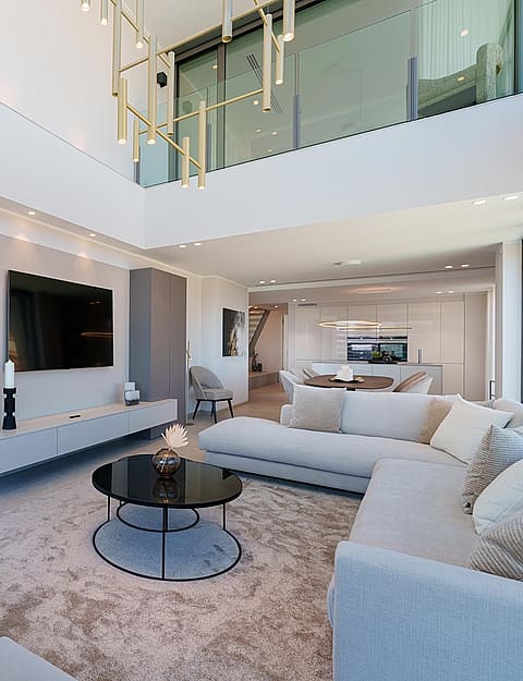 Salón moderno de planta abierta y doble altura con sofá blanco, mesas de cristal y pared equipada para televisión.
