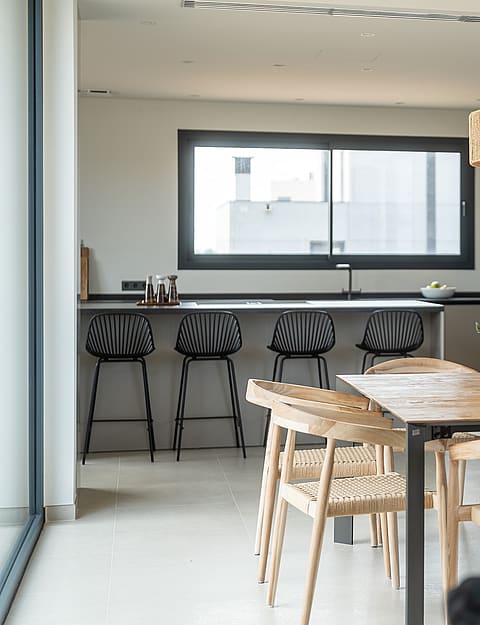 A dining space featuring a large square wooden table with woven chairs, a textured light fixture, and a modern kitchen island with black bar stools in the background.