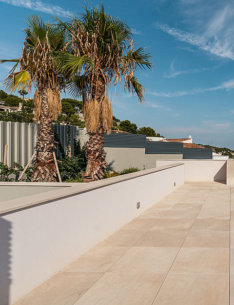 A wide outdoor stone terrace with palm trees, white perimeter walls, and glass railings overlooking a distant coastal landscape.