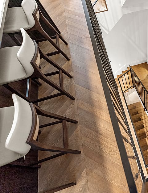 High-angle view of chevron wood flooring, leather-seated bar stools, and a black metal staircase railing.