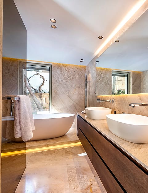 A modern bathroom with dual oval vessel sinks on a wooden vanity, a freestanding white bathtub, and floor-to-ceiling beige travertine stone walls.