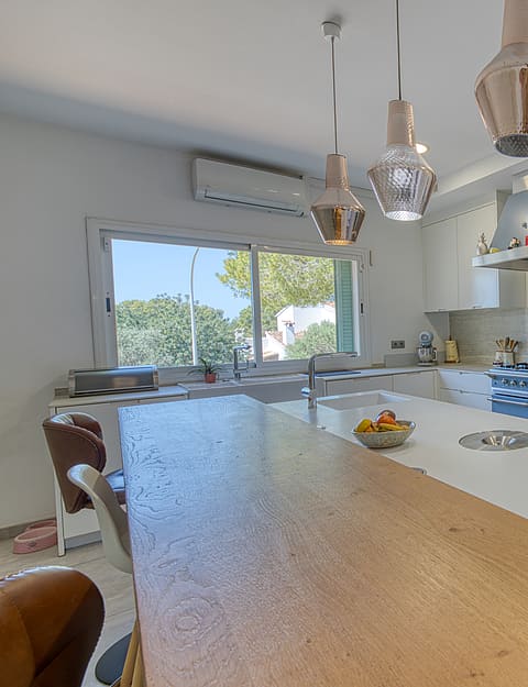 A modern white kitchen featuring a large central island with a wooden breakfast bar, stainless steel stove, and three glass pendant lights hanging above.