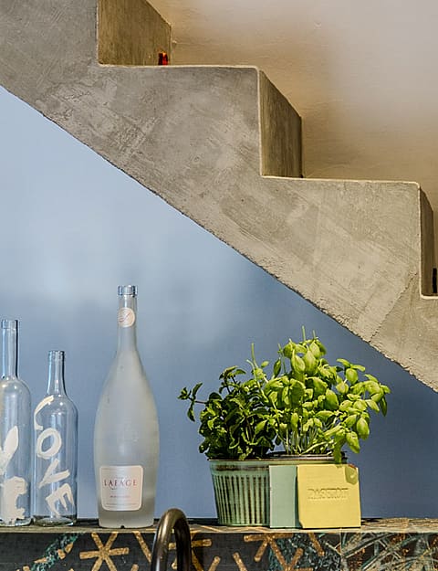 Close-up of a raw concrete staircase with a light blue painted wall, green plant, and glass bottles on a decorative tile ledge.