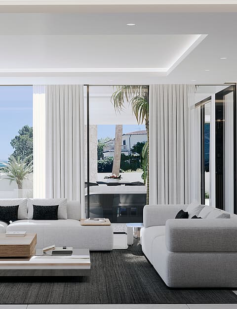 Modern open living room featuring white sofas, a light wood coffee table, dark area rug, and floor-to-ceiling sliding glass doors opening to a covered terrace with coastal views.