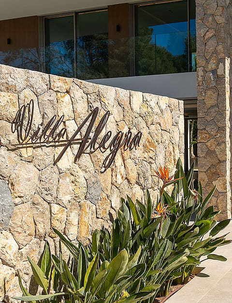 Entrance of a modern villa featuring a dry-stone wall with the name Villa Allegra, stone path, and lush tropical plants.