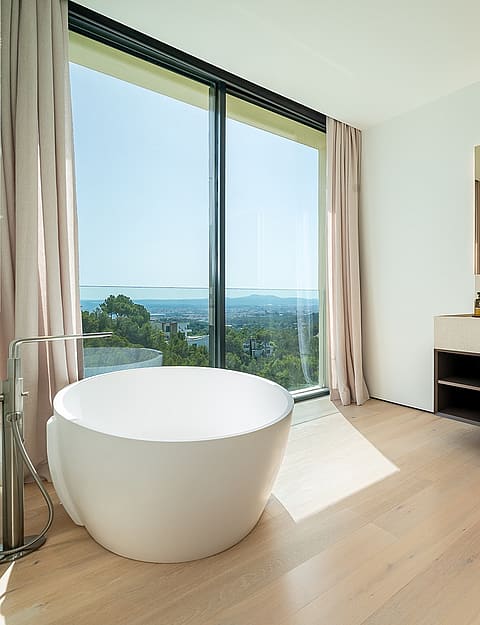 Modern bathroom featuring a white freestanding bathtub positioned beside a large floor-to-ceiling window overlooking the sea.