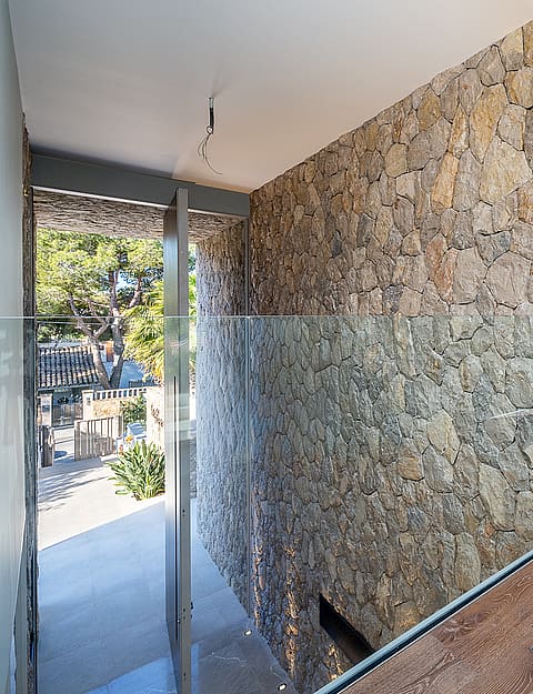 A modern interior hallway features a floor-to-ceiling dry-stone wall, wide oak plank flooring, and a clear glass railing.