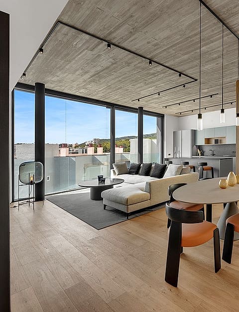 Modern open-plan living and dining room with concrete ceiling, oak flooring, light wood staircase, and floor-to-ceiling windows.