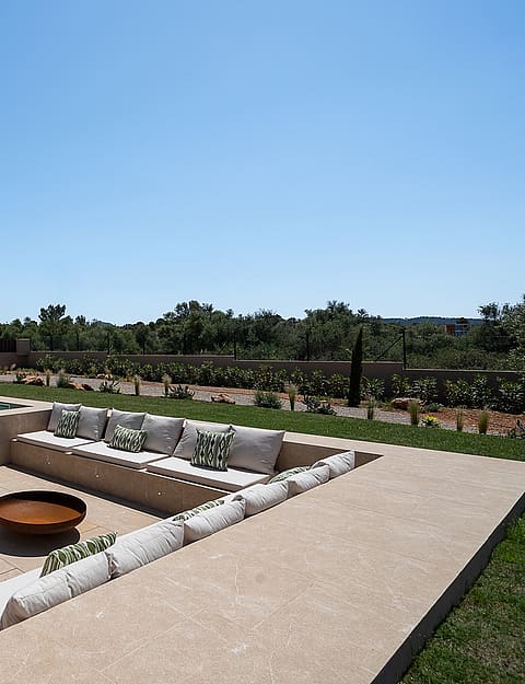 Sunken seating area on a stone terrace featuring built-in benches with cushions and a central fire pit next to a swimming pool.