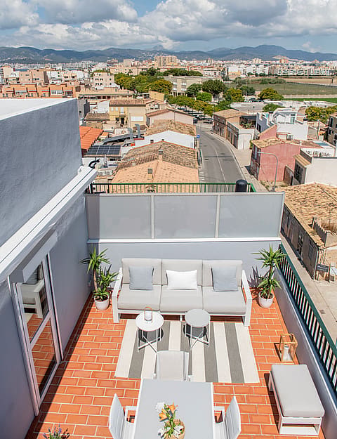 High-angle view of a rooftop terrace with a beige sofa, dining area, and potted plants, overlooking a town and mountains.