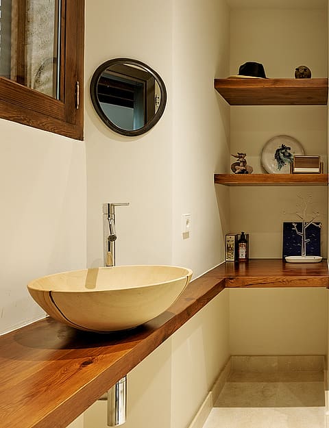 A modern bathroom featuring a circular stone vessel sink on a thick wooden vanity, open shelves, and a walk-in glass shower.