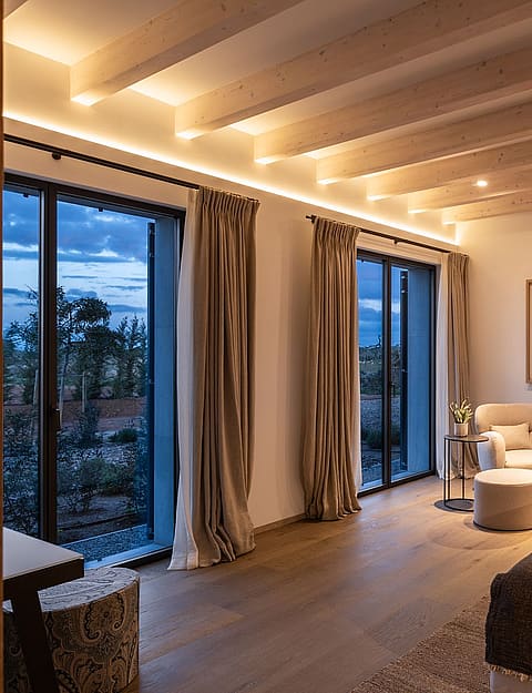 A spacious bedroom with a king-sized bed, exposed wooden ceiling beams, and tall windows showing an evening landscape.