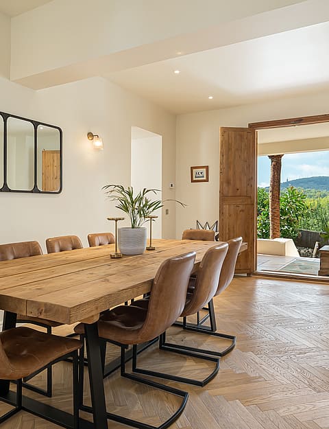 Dining room featuring a long rustic wood table, leather chairs, herringbone flooring, and doors opening to a terrace.