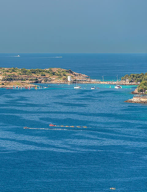 Aerial perspective of the deep blue Mediterranean Sea with a rocky island and distant boats under a clear sky.