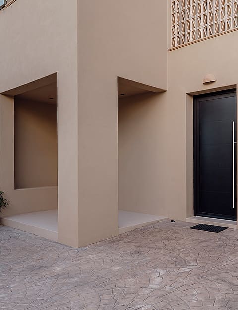 Entrance of a beige townhouse featuring a dark metal front door, gravel pathway, and a decorative concrete block screen.