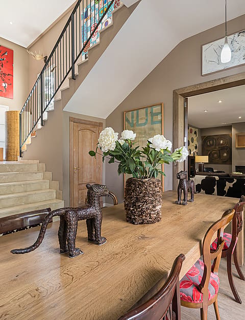 Interior view of an entryway with a stone staircase, wooden dining table, and large floral arrangement, looking through to a living room.