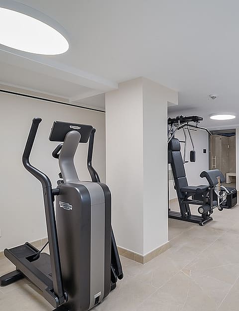Interior de gimnasio doméstico con elíptica, máquina de musculación y sauna acristalada.