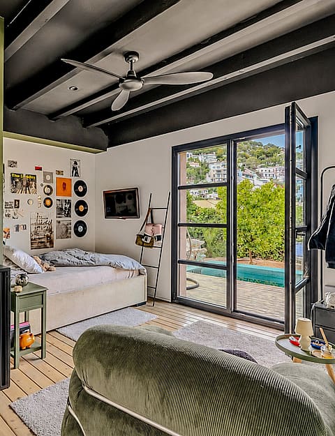 A bedroom with sage green walls, dark ceiling beams, a ceiling fan, and black framed glass doors opening to a garden patio.