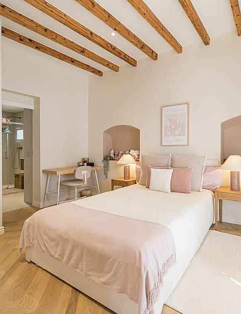 Bright bedroom featuring exposed ceiling beams, herringbone wood floors, and recessed wall arches above a double bed.