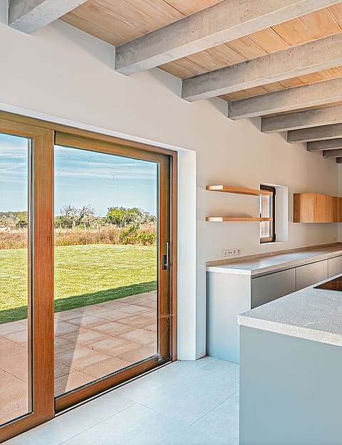 Modern kitchen featuring a central island, light grey cabinetry, wooden ceiling beams, and large glass doors overlooking a field.
