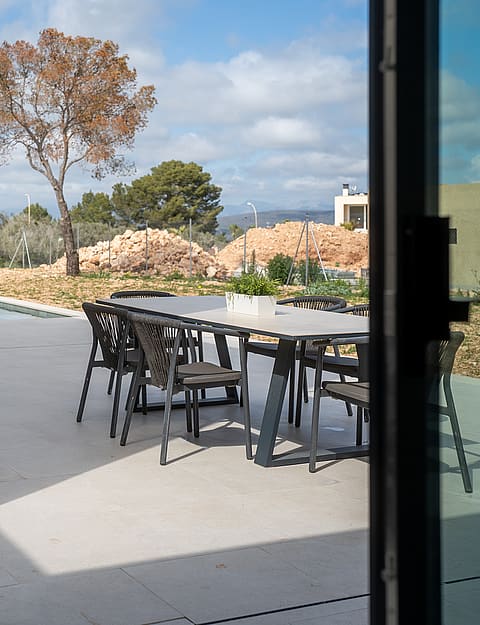 A covered terrace with a dining set and sun loungers overlooks a swimming pool and distant hills under a sunny sky.
