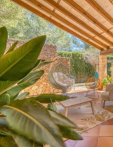 A covered terrace with terracotta floor tiles and wooden beam ceiling, featuring outdoor armchairs and a dry stone wall.
