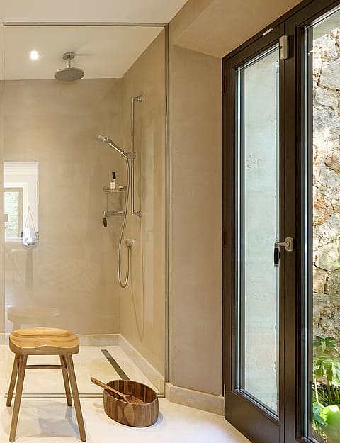 A minimalist bathroom with beige walls, a walk-in glass shower, wall-mounted vanity, and a wooden stool, featuring a glass door leading outside.