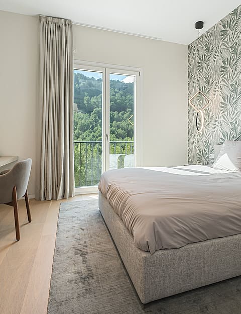 A modern bedroom featuring a patterned leaf-print wallpaper, a platform bed with grey bedding, and a built-in desk area.