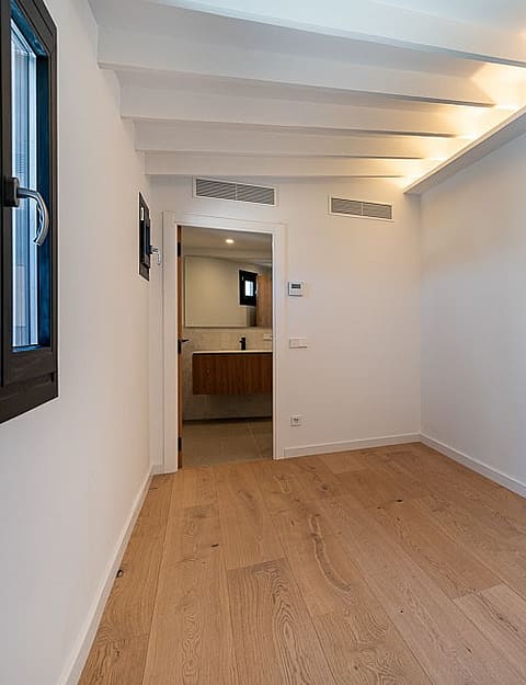 A minimalist bedroom interior featuring light wood flooring, white walls with beamed ceilings, and a tall integrated wardrobe.